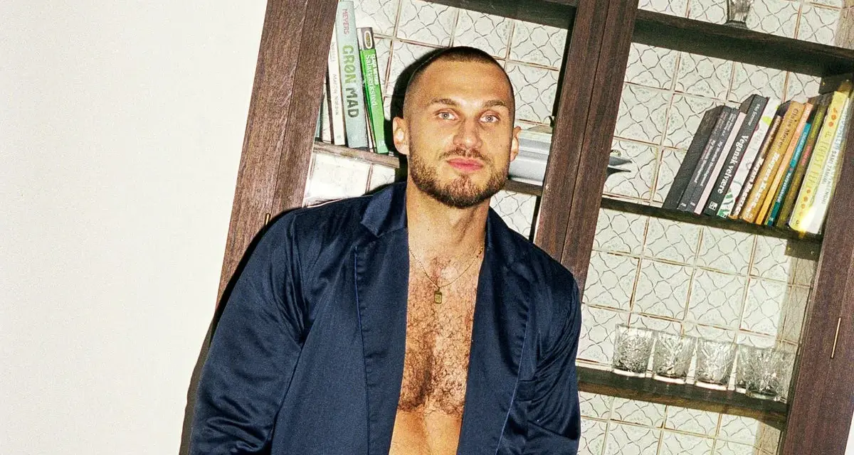 Hombre con corte afeitado, barba y chaqueta abierta, posando frente a una estantería con libros y vasos.