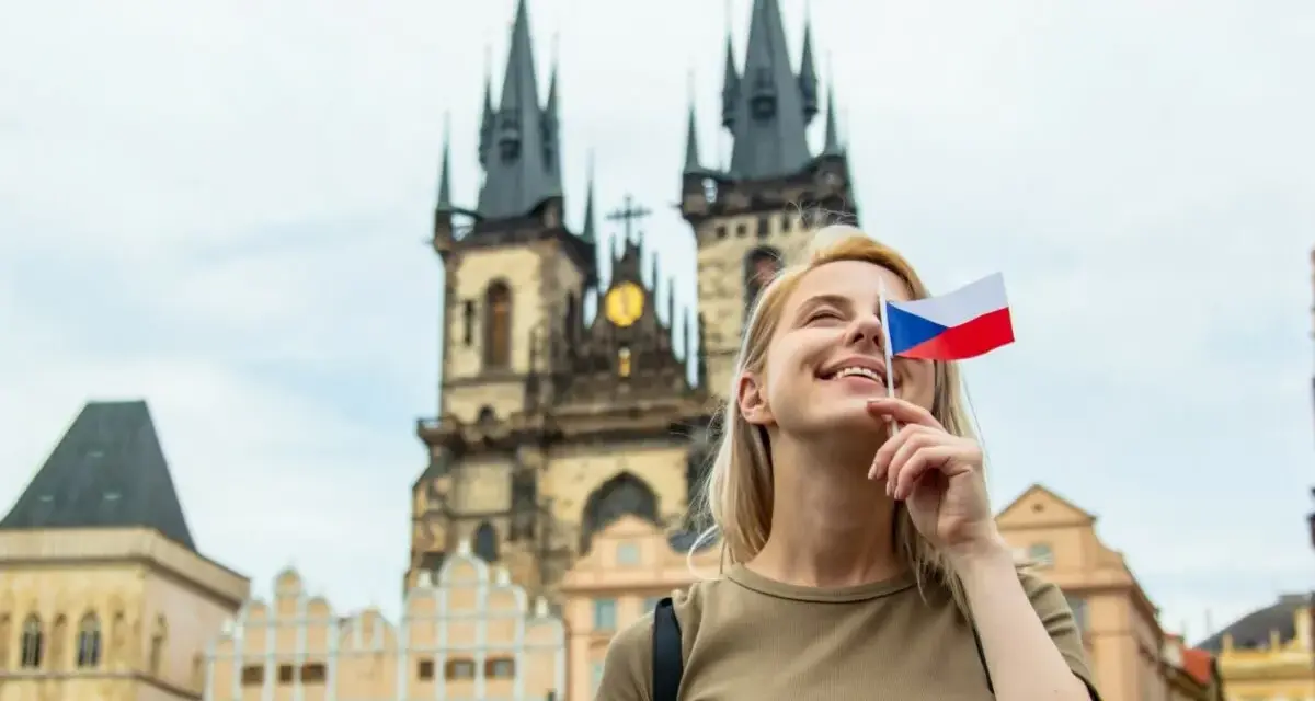 Uśmiechnięta blondynka z flagą Czech zasłaniającą oko, marzy o wysokich zarobkach w Czechach. W tle praskie kamienice i kościół.