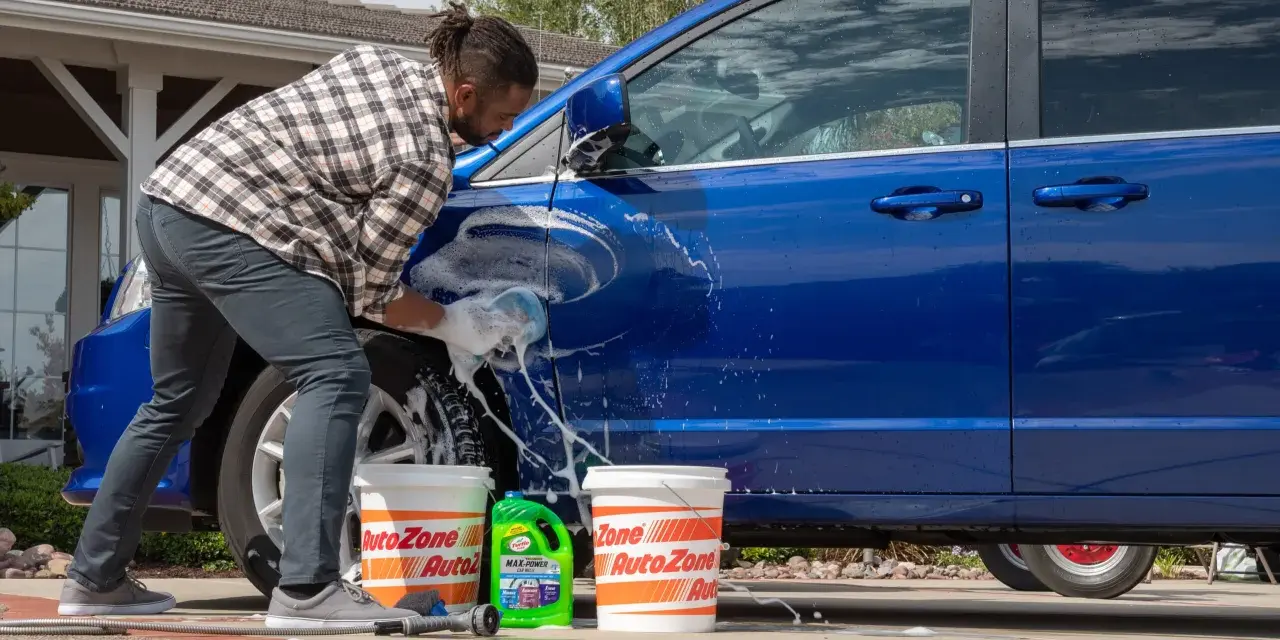 A man washes a blue car in the sun, with buckets and car wash soap nearby. It's a good day to wax a car in the sun.