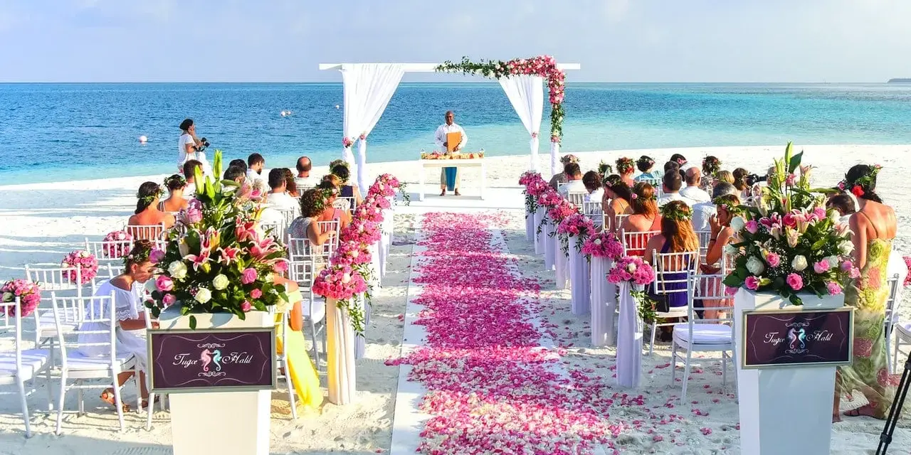 Piękna plażowa ceremonia ślubna. Kwiaty i płatki róż tworzą romantyczną ścieżkę. Goście siedzą na białych krzesłach, a ocean w tle.