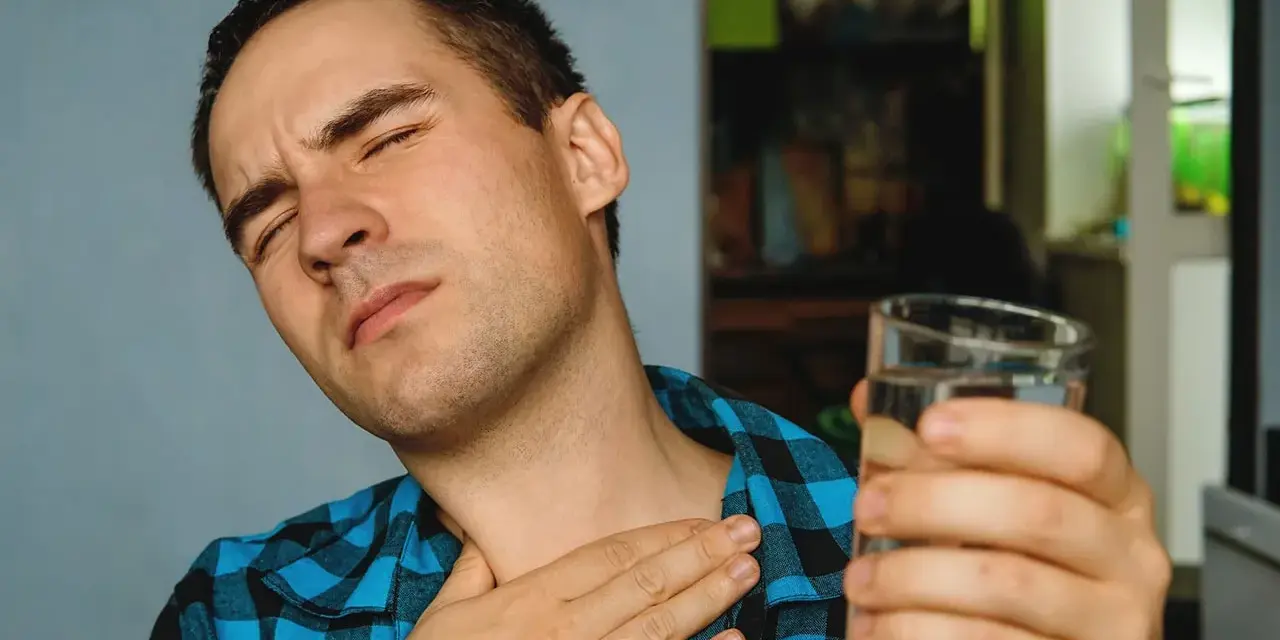 Mężczyzna z bólem gardła, cierpiący na nocny kaszel po alkoholu, sięga po szklankę wody.