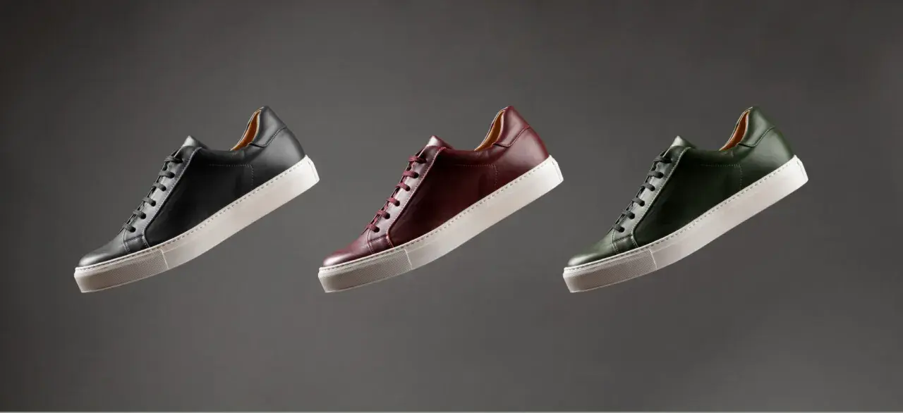 Sneakers marche famose in pelle nera, bordeaux e verde, con suola bianca, fluttuano su sfondo grigio.