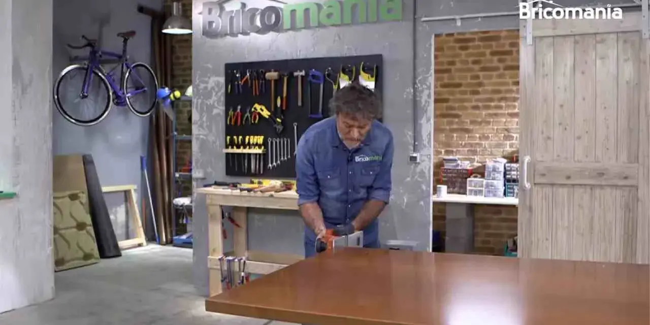 Hombre lijando mesa de madera. El taller tiene herramientas colgadas y una bicicleta morada. La madera hinchada por humedad se repara.