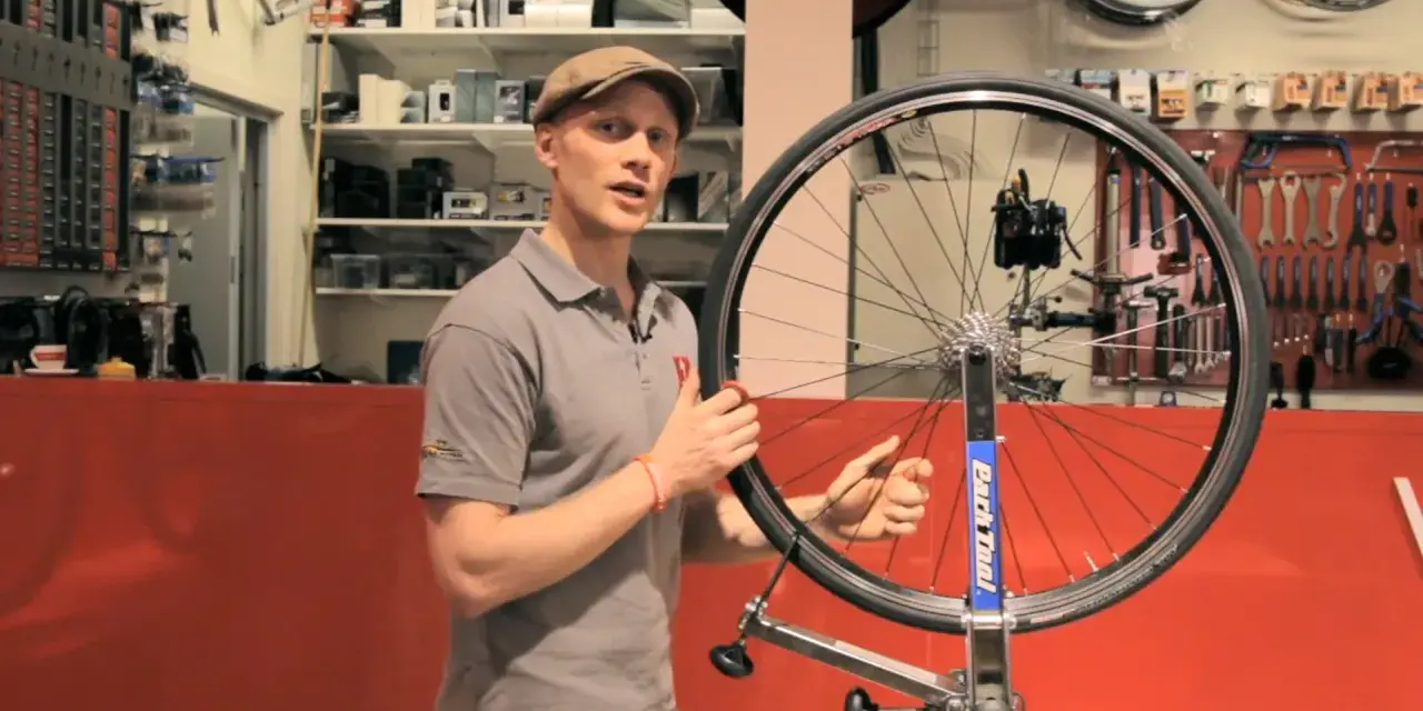 Man i keps visar hur man rikta cykelhjul i en verkstad. Verktyg hänger på väggen bakom.
