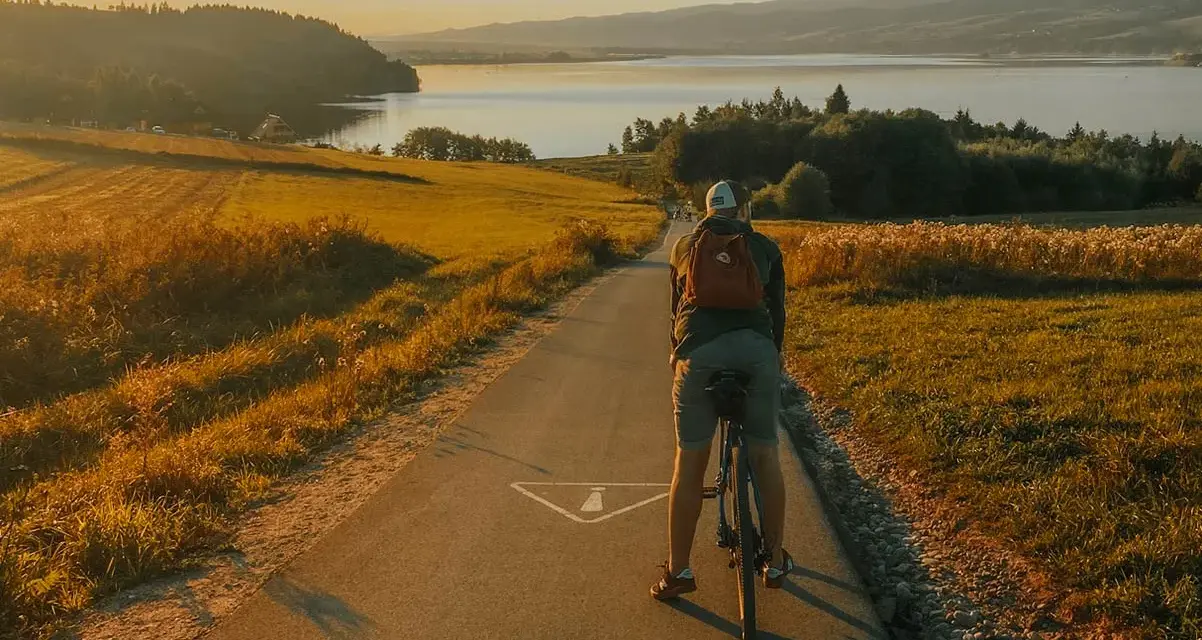 Rowerzysta na ścieżce rowerowej w kierunku jeziora. Złote światło zachodu słońca oświetla malowniczą trasę Velo Czorsztyn.