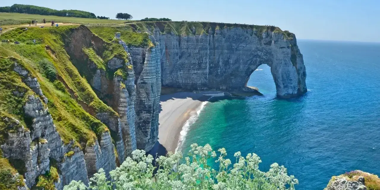 Falaises d'Étretat, France. L'arche naturelle de la Porte d'Aval se dresse fièrement au-dessus de la plage de galets. Le **gr 21** serpente le long de la côte verdoyante.