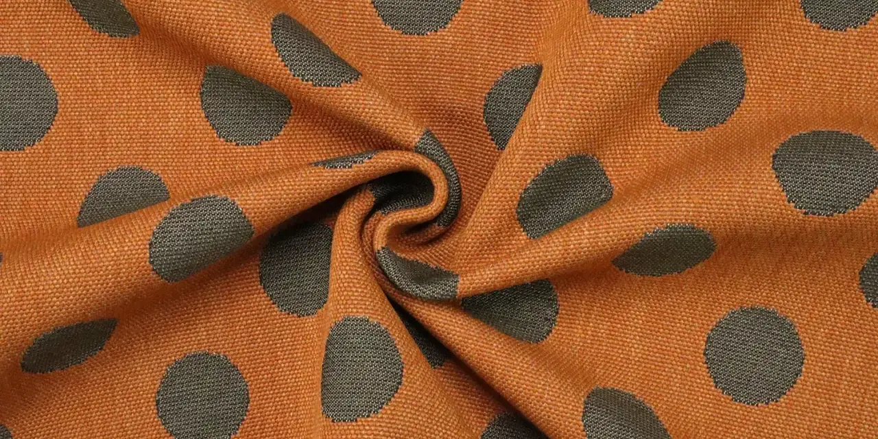 Orangefarbener Stoff mit dunklen Punkten, die wie kleine Kreise aussehen. Das Muster ist unregelmäßig und wirkt lebendig.
