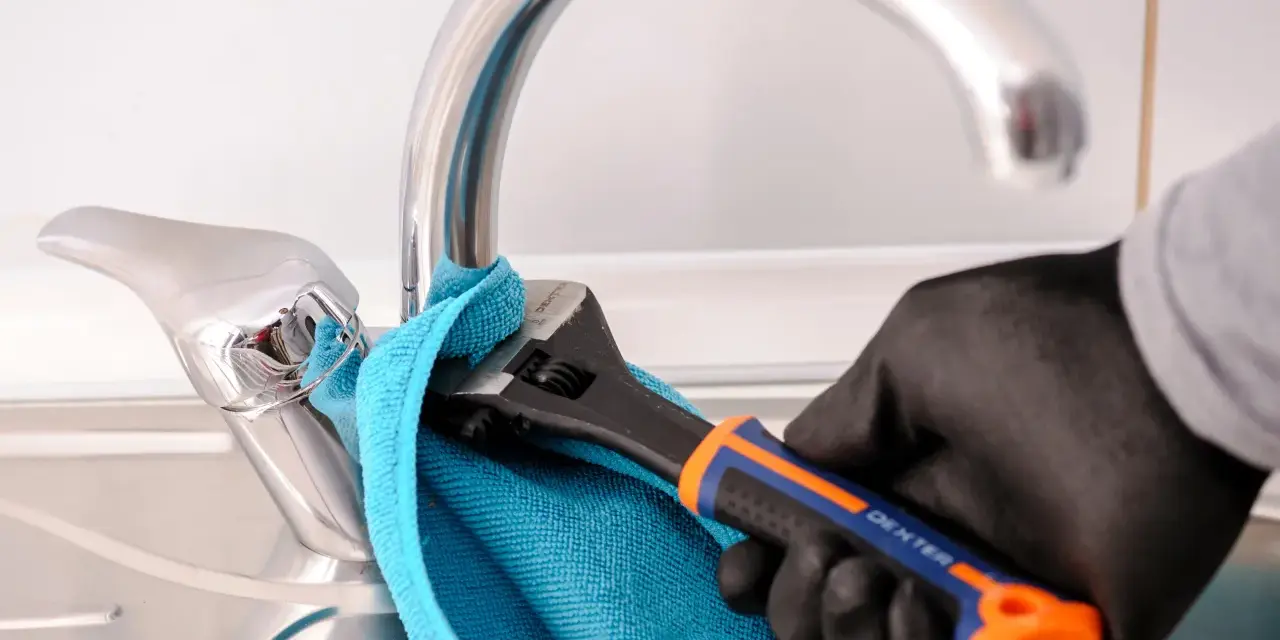 Mano enguantada usa llave inglesa para cambiar grifo cocina. Toalla azul protege el cromo.