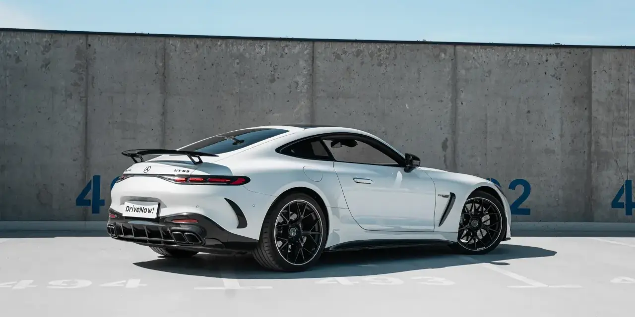 Biały Mercedes GT 63 z czarnym spojlerem i felgami, zaparkowany pod betonową ścianą z niebieskimi cyframi.