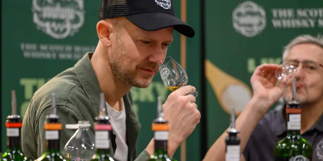 Mann mit Kappe riecht an einem Whiskyglas bei einer Whisky Messe. Flaschen stehen auf dem Tisch.