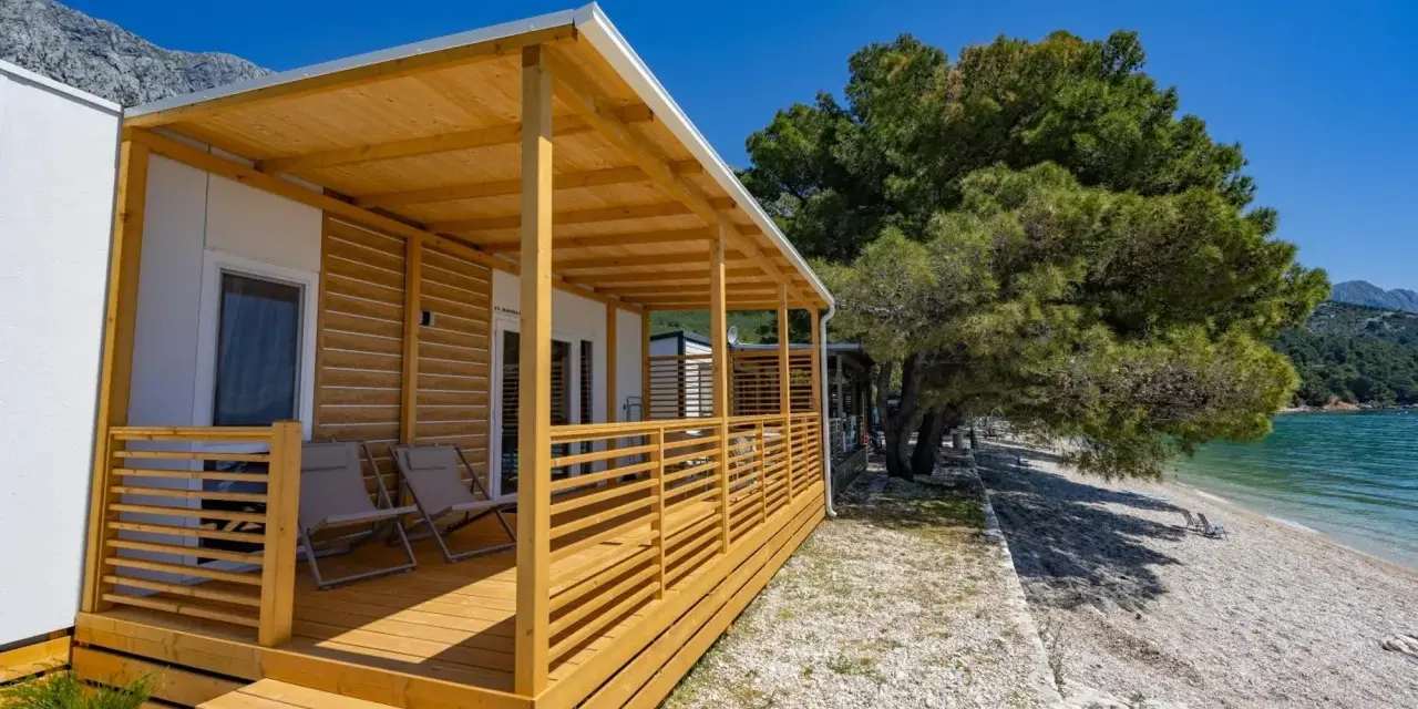 Domek mobilny z drewnianym tarasem i widokiem na morze w camp Dole Živogošće. Dwa leżaki czekają na plaży.