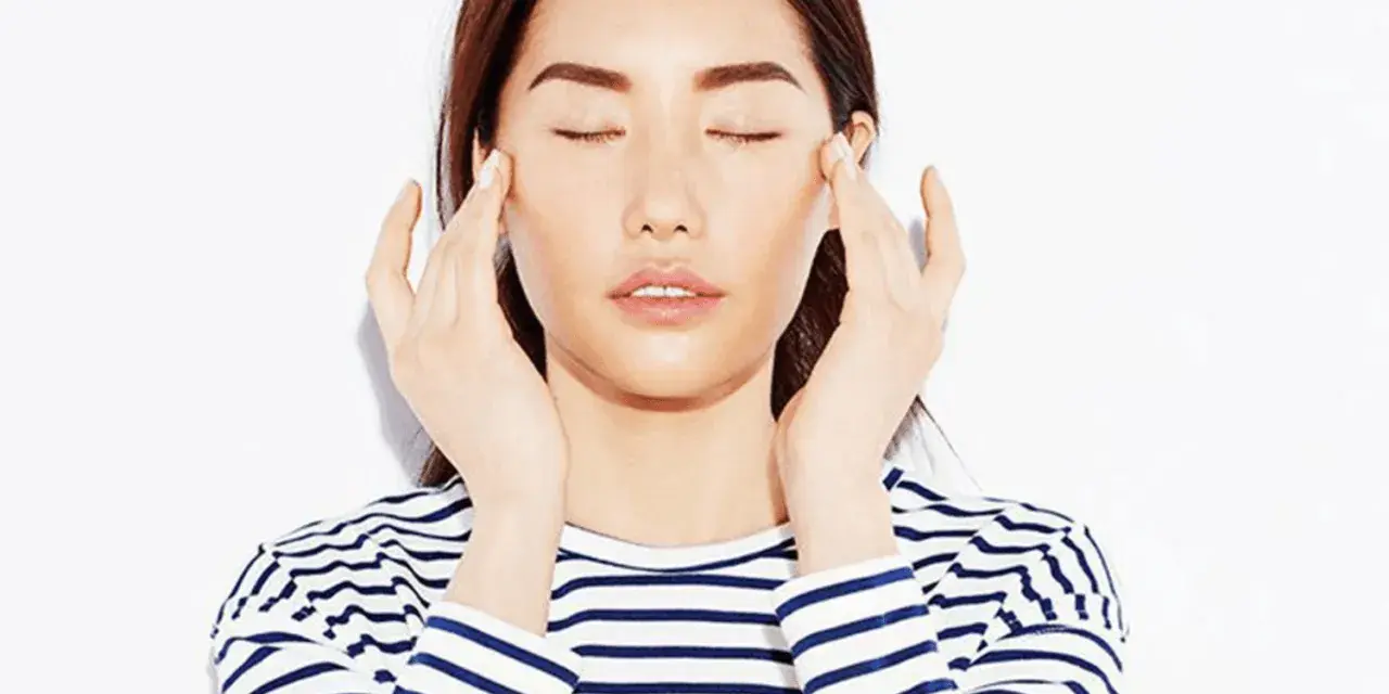 A woman with closed eyes performs a gentle face massage for puffiness, her fingers delicately touching her temples.