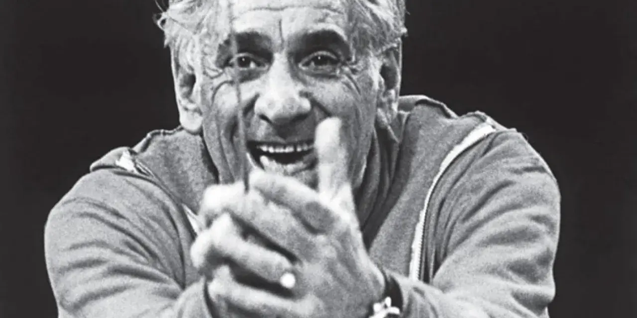 Uśmiechnięty Leonard Bernstein, dyrygent, z dłońmi złożonymi przed sobą, wpatruje się w widza.