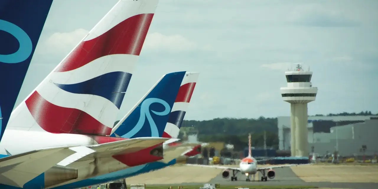 Samoloty z charakterystycznymi ogonami British Airways i innym logo czekają na start na lotnisku Londyn Gatwick. W tle wieża kontroli lotów.