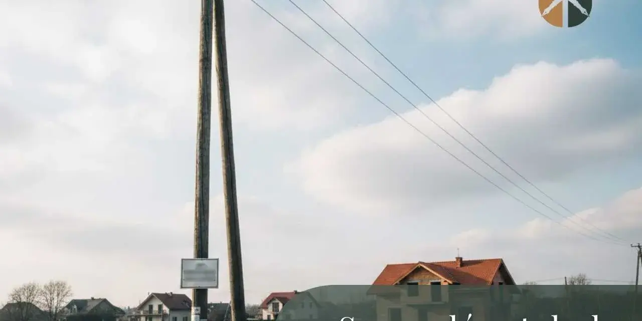 Drewniany słup z przewodami tworzy linię napowietrzną nad polami i domami.