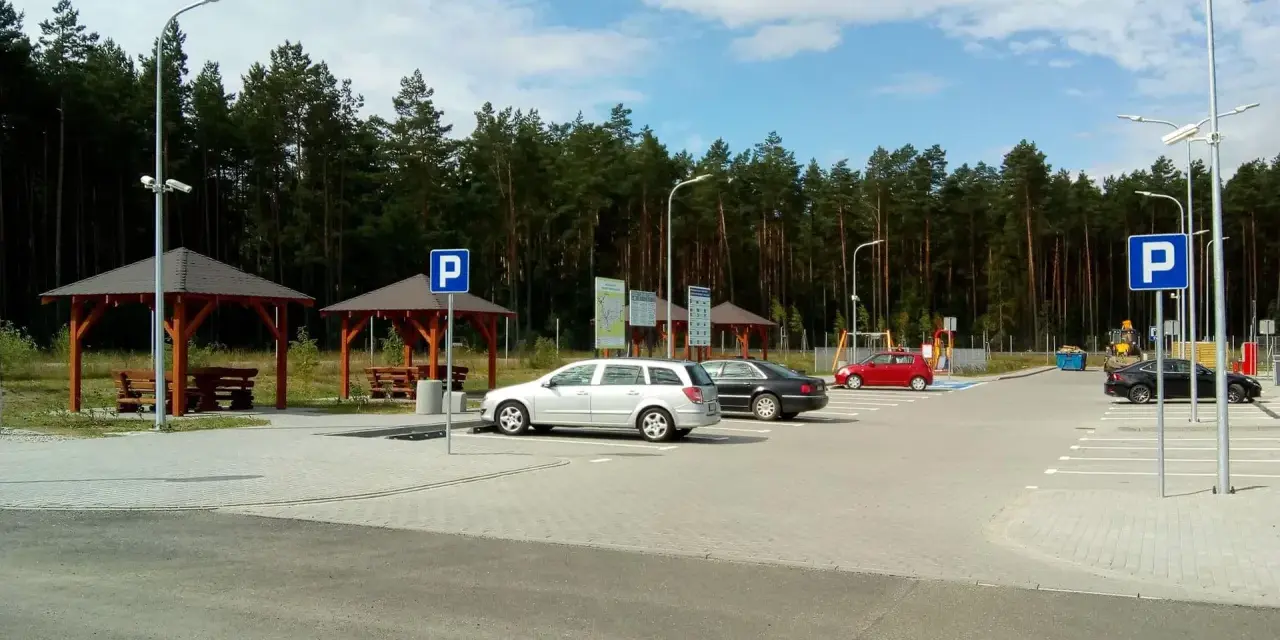 Parking przy lesie, z wiatami i ławkami. W tle las, a na parkingu kilka samochodów. To idealne miejsce na postój w drodze, może nawet w pobliżu **Orlen Sople**.
