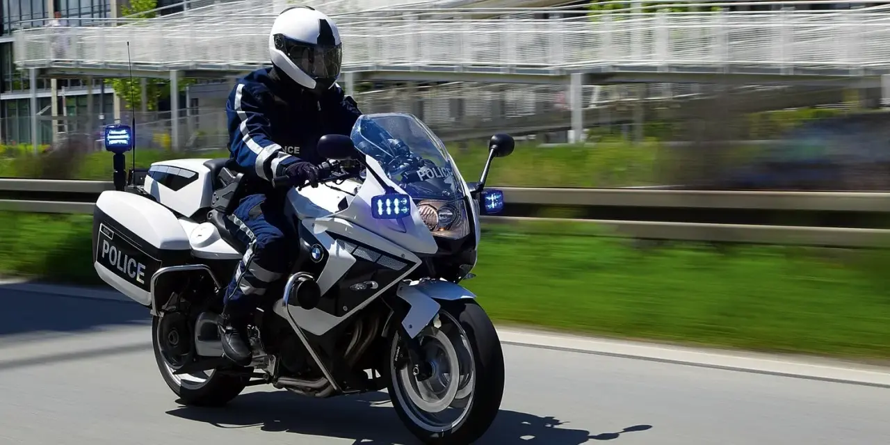 Policyjny motocykl BMW F800GT z włączonymi sygnałami świetlnymi pędzi po drodze.