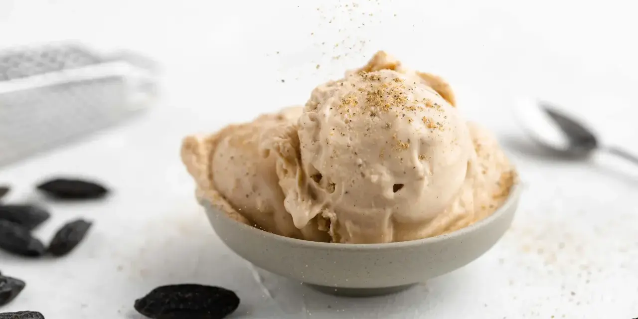 Cremiges Tonkabohnen Eis in einer Schale, bestreut mit gemahlenen Tonkabohnen. Mehrere ganze Tonkabohnen liegen daneben.