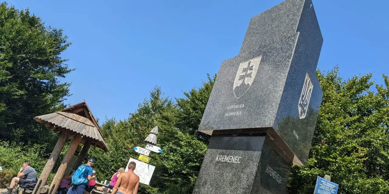 Trójstyk granic Bieszczady: monumentalny obelisk z herbami Słowacji i Ukrainy, obok drewniana wiata i turyści.