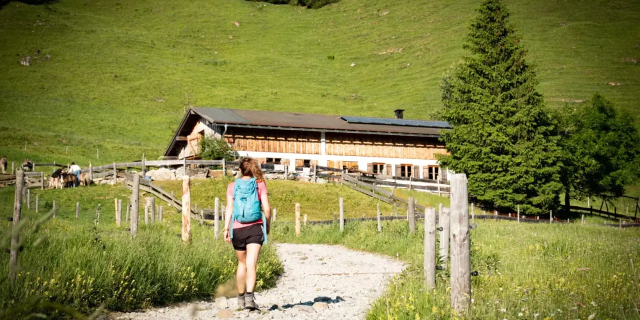 Frau mit Rucksack wandert auf einem Schotterweg zu einer Berghütte. Grüne Wiesen und Bäume im Hintergrund.