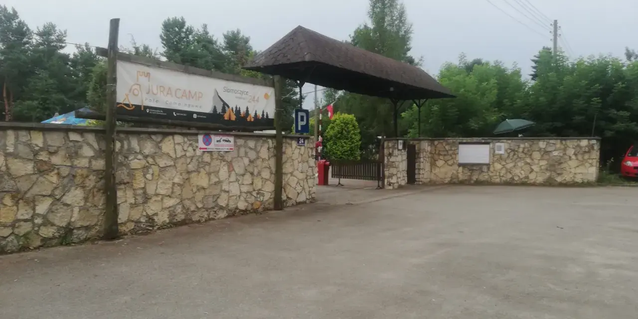 Wjazd na siamoszyce camping przez kamienny mur. Baner "Jura Camp" zaprasza do wypoczynku wśród drzew.