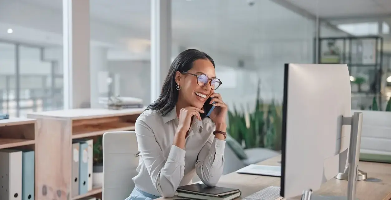 Donna sorridente al telefono in ufficio, mentre lavora al computer. Potrebbe chiedersi quanto guadagna una segretaria al mese.