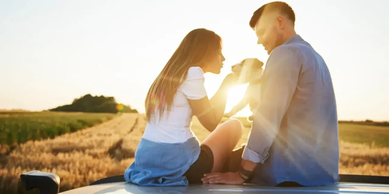 Ein Paar sitzt auf einem Auto im Sonnenuntergang, ein Hund zwischen ihnen. Sie scheinen in einer stillen Partner Meditation zu sein, umgeben von Feldern.