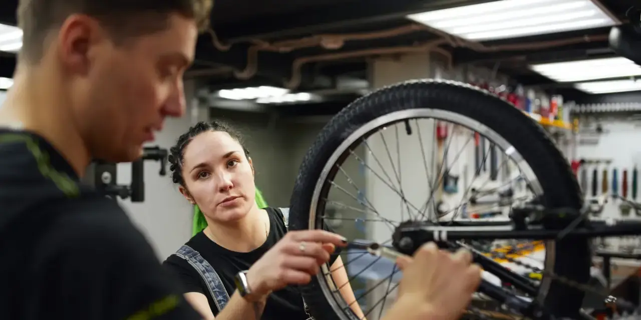 Mechanik rowerowy wyjaśnia, ile kosztuje wymiana dętki w rowerze, podczas gdy kobieta z zielonymi włosami uważnie słucha.
