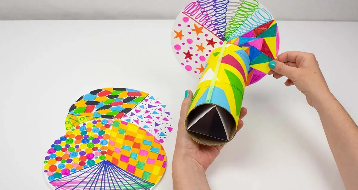 Manos creando manualidades con material reciclado para adolescentes: un caleidoscopio con tubos de cartón y discos decorados con patrones coloridos.
