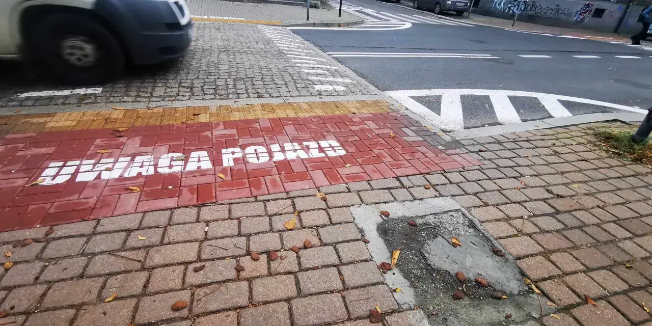 Czerwony pas na chodniku z napisem "UWAGA POJAZDY" informuje o przejściu sugerowanym. W tle widać samochody i schody.