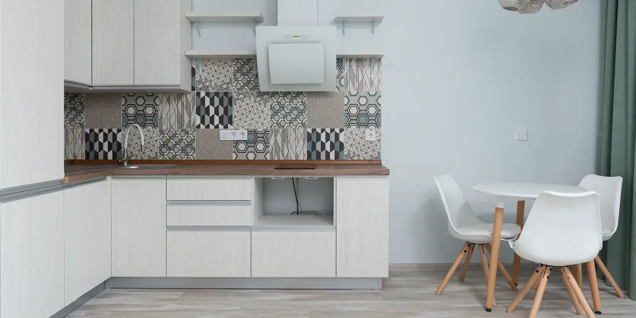 Cocina moderna con armarios blancos, salpicadero geométrico y suelo gris claro. Mesa redonda con sillas blancas.