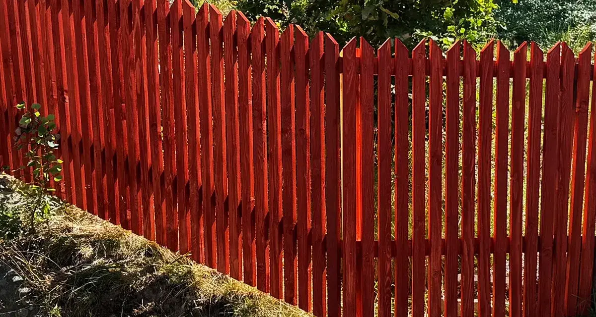 Rött trästaket med spetsiga toppar ger fin staket inspiration. Grönskande träd i bakgrunden.