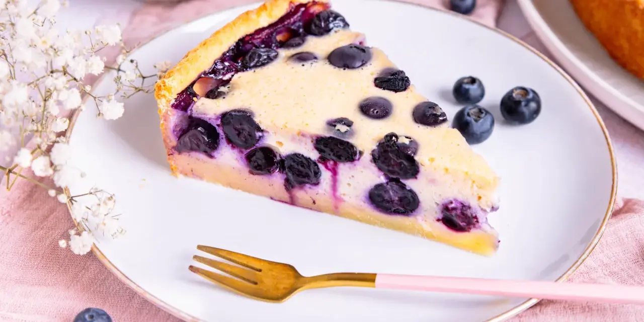 Ein Stück Heidelbeerkuchen Landfrauenart mit saftigen Blaubeeren und cremigem Guss auf einem Teller, daneben eine Gabel und frische Beeren.