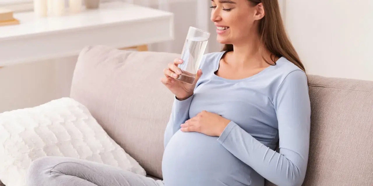 Uśmiechnięta ciężarna kobieta pije wodę, dbając o siebie podczas ciąży. Krzywa cukrowa w ciąży wymaga troski.
