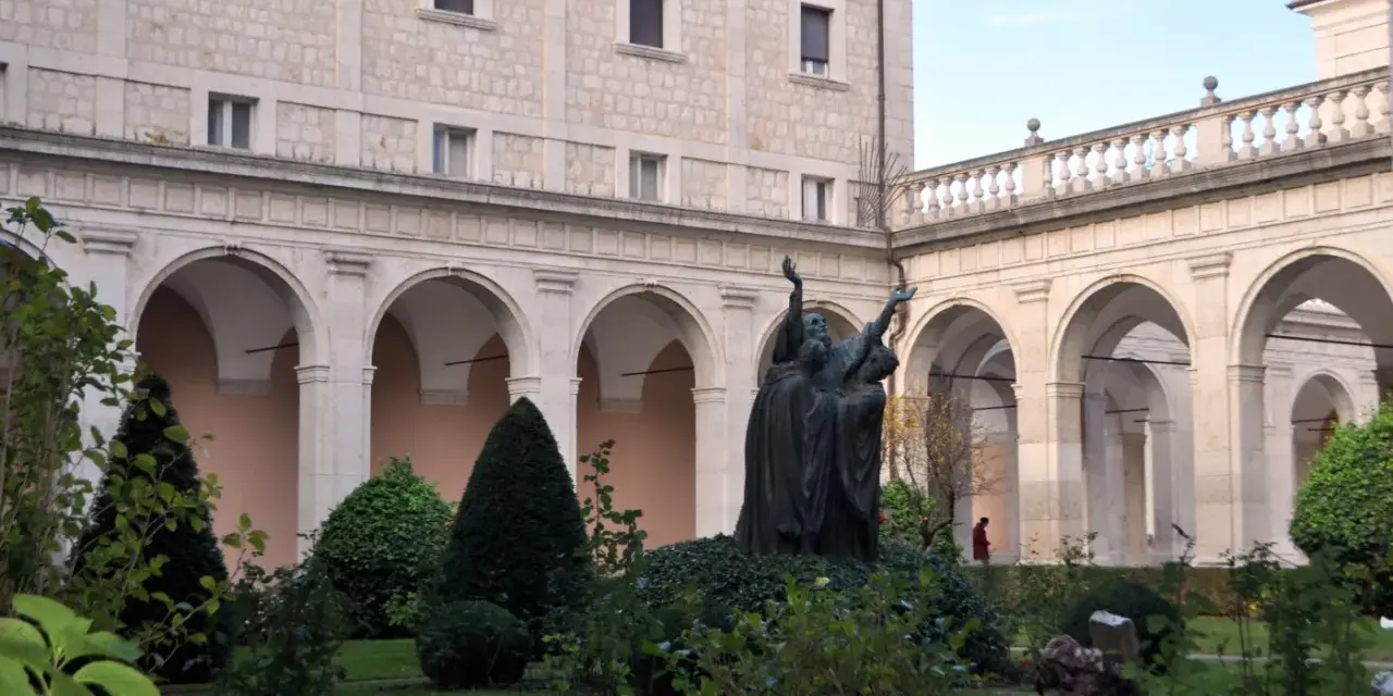 A bronze statue of monks with arms raised stands in a courtyard garden, perhaps commemorating St. Benedict's feast day.
