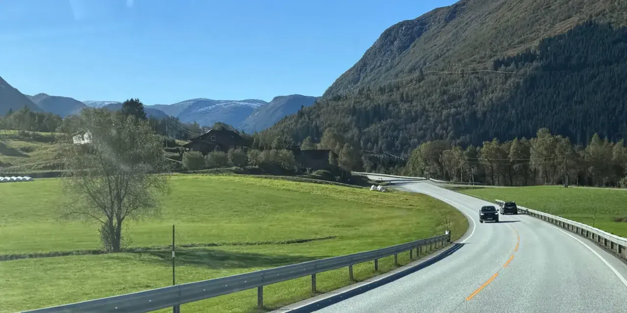 En bil kör på en slingrande väg genom ett grönt landskap med berg och hus. En perfekt start på en semester i Norge.