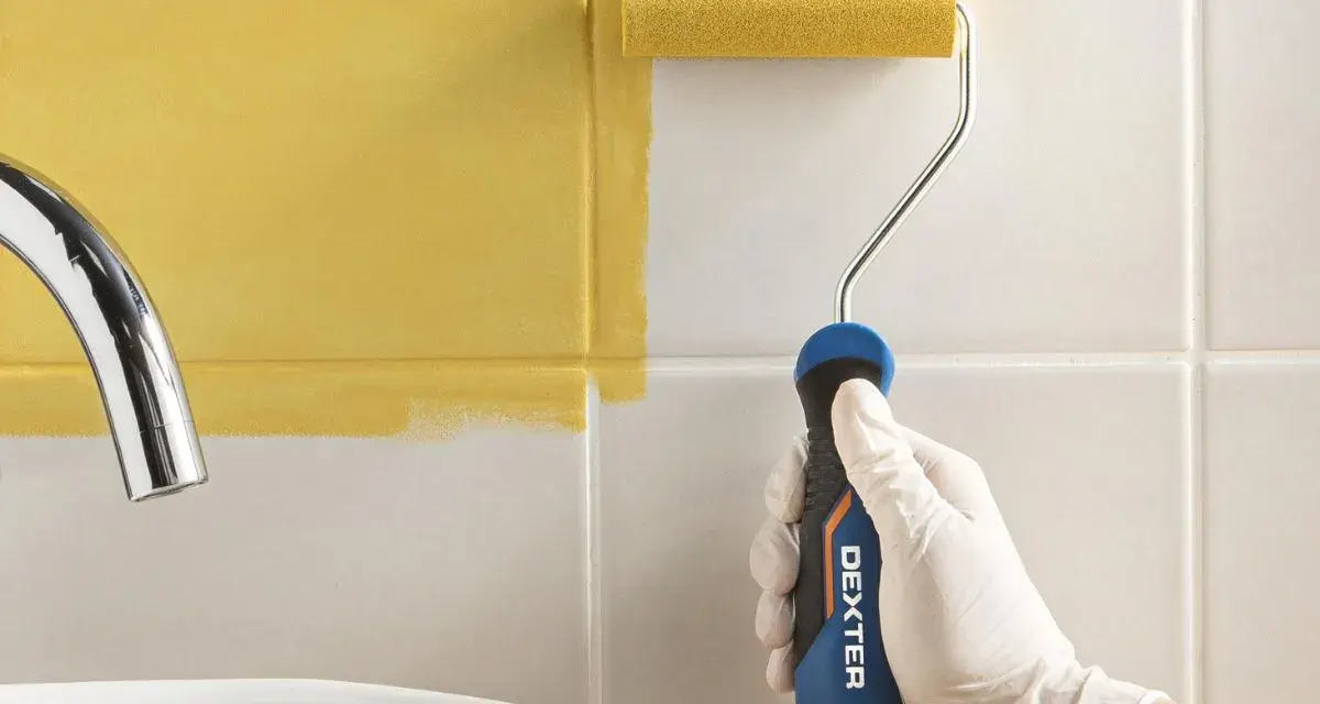 Mano con guante blanco pintando azulejos blancos con rodillo amarillo.