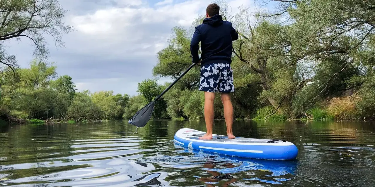 Mann auf SUP-Tour auf einem ruhigen Fluss, umgeben von üppiger Vegetation.