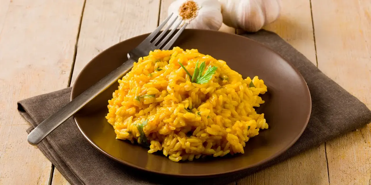 Un invitante risotto zafferano in polvere, guarnito con prezzemolo fresco, su un piatto marrone, con aglio e forchetta sullo sfondo.