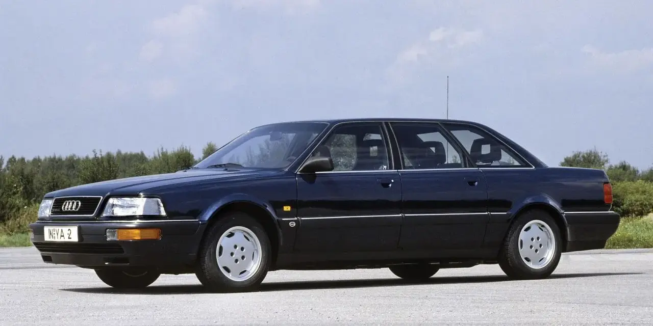 Ciemnoniebieski Audi V8 z lat 90. stoi na pustej drodze, z drzewami w tle.