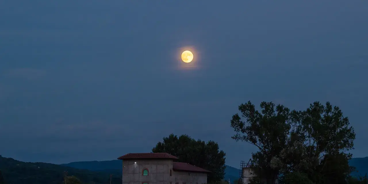 La luna di luglio illumina un paesaggio serale con colline, alberi e un edificio.