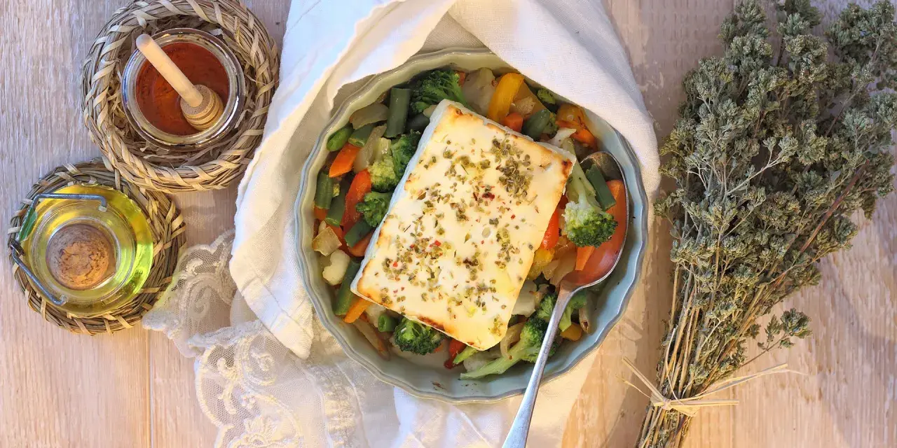 Feta al forno con verdure miste, condita con erbe aromatiche. Accanto, olio, spezie e un mazzetto di origano secco.