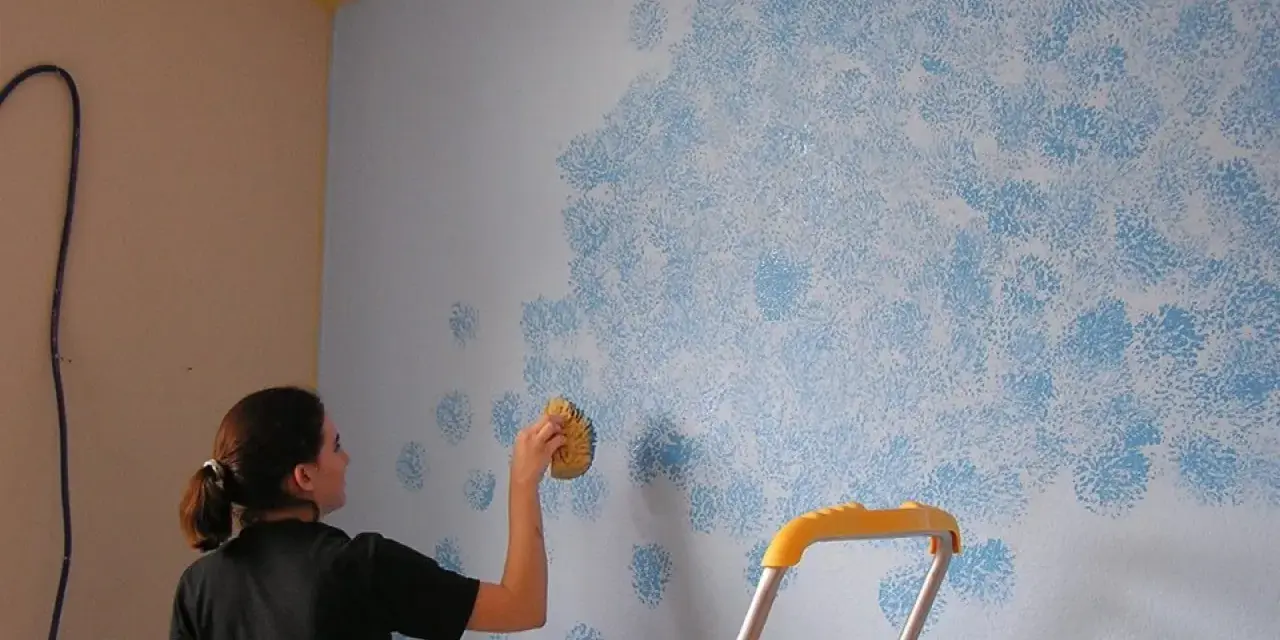 Mujer aplica pintura que disimula imperfecciones en pared con esponja, creando un patrón de nubes azules.