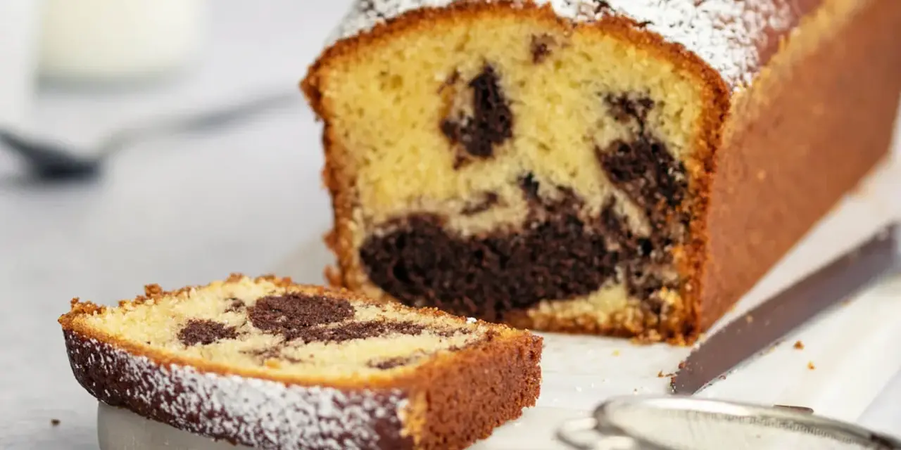 Ein saftiger Marmorkuchen in Kastenform, bestäubt mit Puderzucker. Eine Scheibe liegt daneben, bereit zum Genuss.