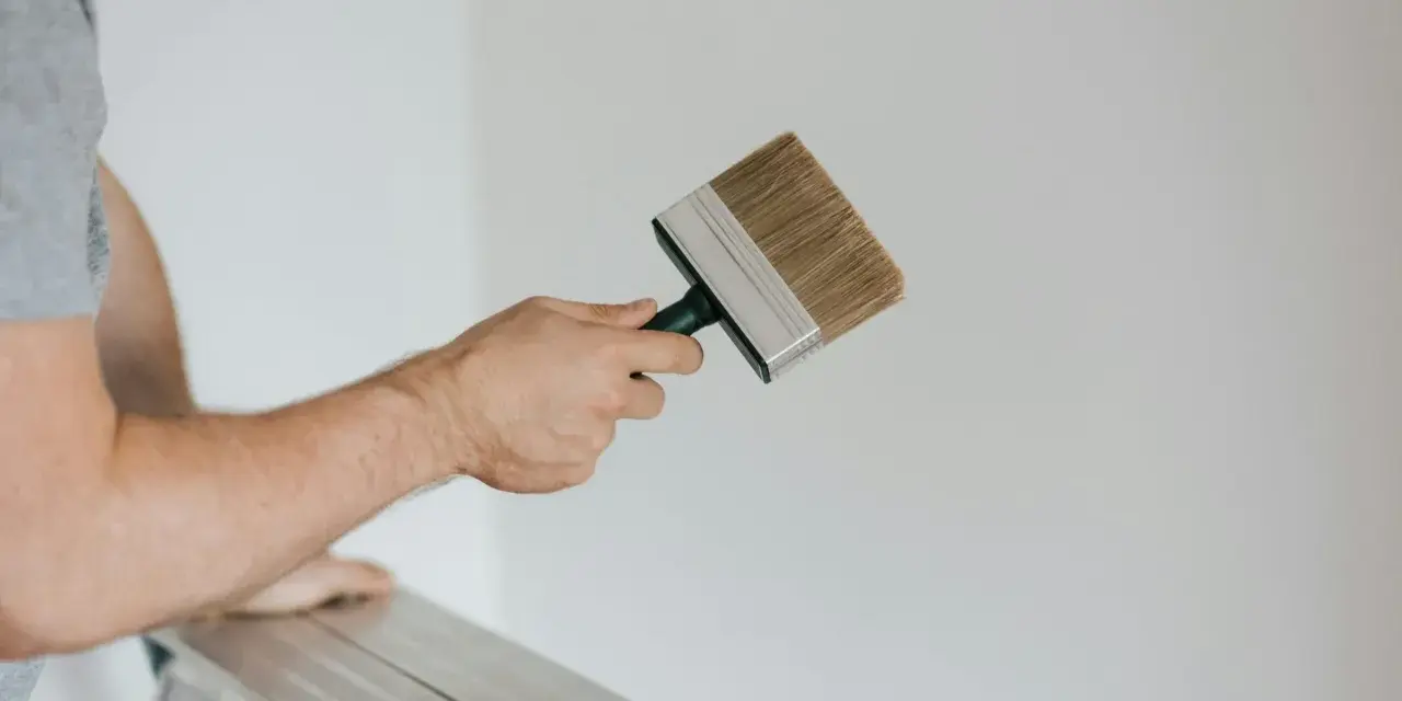 Person på en stege håller en bred pensel redo att måla huset vitt.