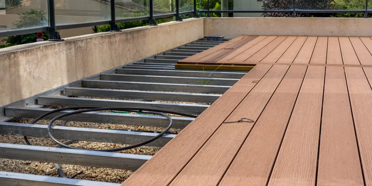 WPC Terrasse Ideen: Unterkonstruktion aus Metall mit Kiesbett und beginnender Verlegung von braunen WPC-Dielen. Moderne Glasbrüstung.