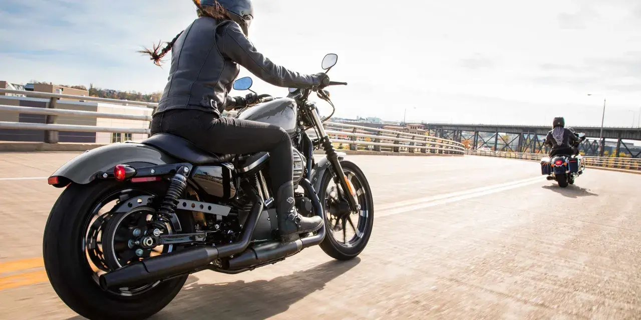 Kobieta w skórzanej kurtce i kasku jedzie na stylowym motocyklu dla kobiety. Wiatr rozwiewa jej włosy, a słońce odbija się od chromowanych elementów.