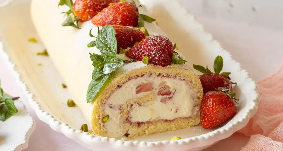 Eine köstliche Erdbeer Biskuitrolle, verziert mit frischen Erdbeeren, Minze und Pistazien, auf einem weißen Teller.