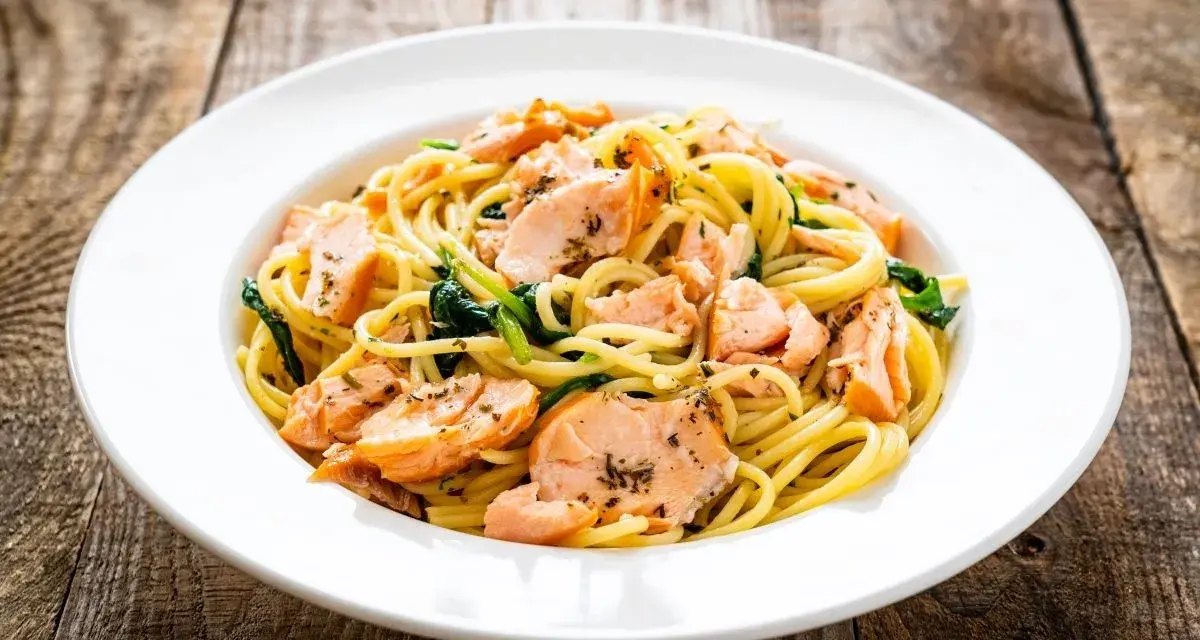Makaron spaghetti z łososiem i szpinakiem na białym talerzu, na drewnianym tle.