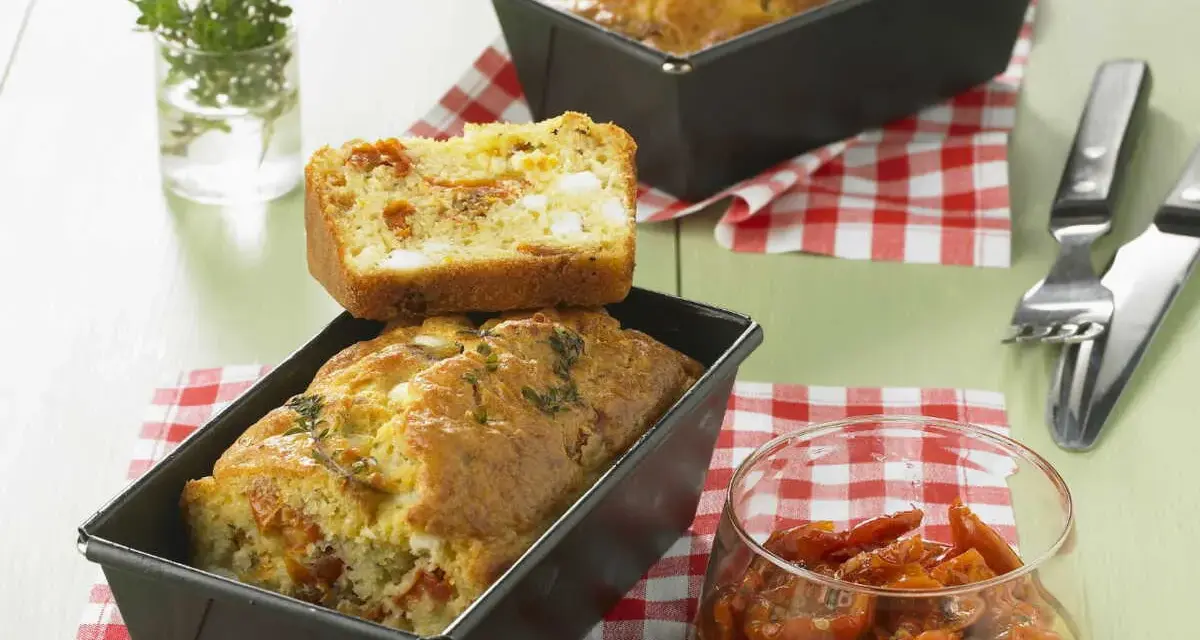 Cake tomates séchées feta, un délice salé aux herbes fraîches. Une tranche est posée sur un cake encore dans son moule.
